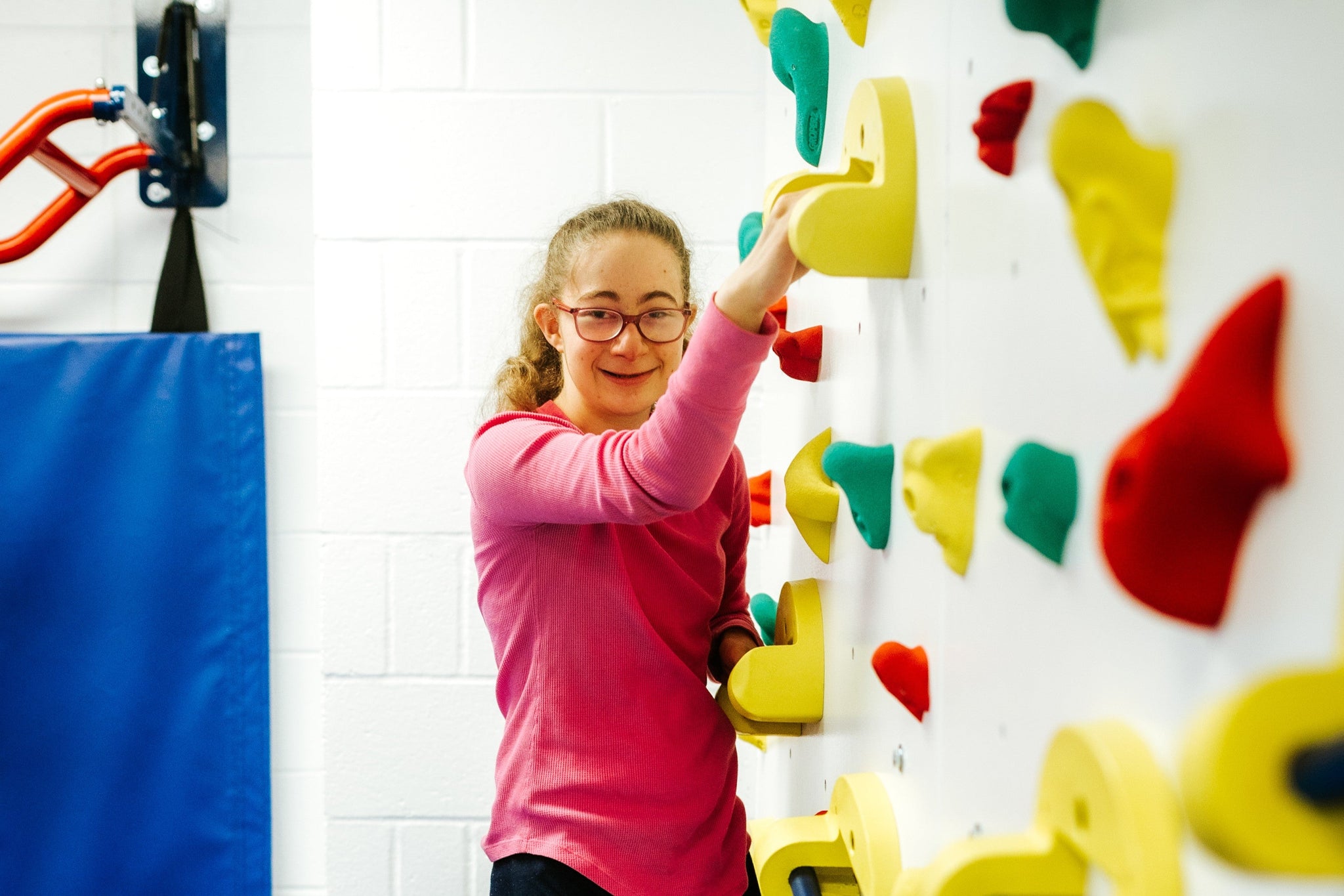 this action room features a colorful indoor climbing wall, complete with safety crash mats and a smiling girl holding the climbing handholds.