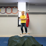 child balancing while standing on bottom bar of a double trapeze bar swing indoors