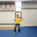 child sitting and swinging indoors from bottom bar of a double trapeze bar swing.