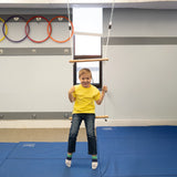 child sitting and swinging indoors from bottom bar of a double trapeze bar swing.